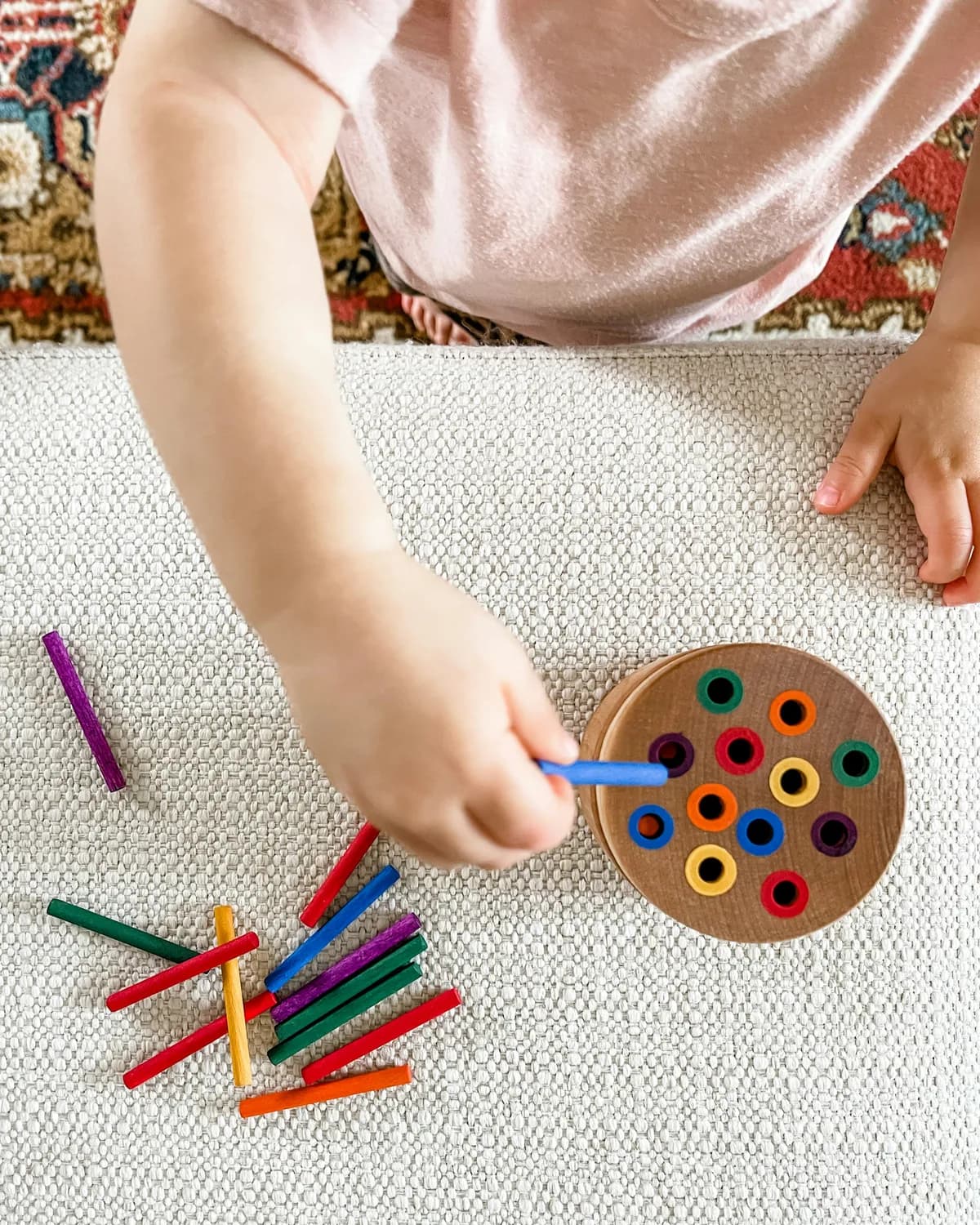 A child's hand placing a coloured peg into a wooden sorting disc on a woven rug